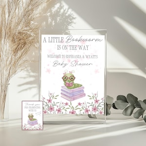 Puede incluir: Un letrero de baby shower con el texto "A Little Bookworm Is On The Way" y "Welcome to Esperanza & Wyatt's Baby Shower." El letrero presenta un gusano de libro de dibujos animados leyendo un libro, con flores rosas.