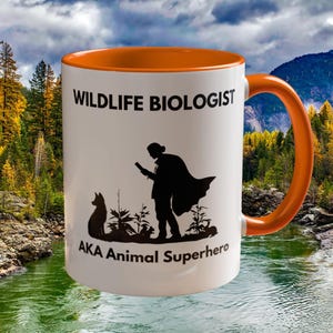 May include: White ceramic mug with an orange interior and handle. The mug features the text "WILDLIFE BIOLOGIST" above a silhouette of a person with a cape and a dog, with the text "AKA Animal Superhero" below.