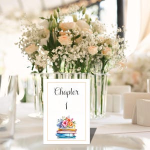 May include: A white table card with gold trim features the word "Chapter" above the number "1". Below, a watercolor illustration depicts a stack of books topped with colorful flowers. A vase of flowers is in the background.