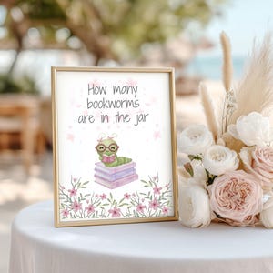 May include: A framed sign with the text "How many bookworms are in the jar" features a cartoon bookworm wearing glasses and reading a book atop a stack of books. The sign is decorated with pink flowers and is set on a white table with a floral arrangement.