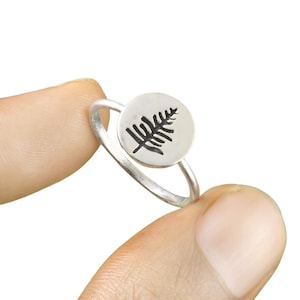 Puede incluir: Un anillo de plata con un diseño de helecho redondo. El helecho es negro y el anillo es plateado.