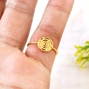 Puede incluir: Un anillo dorado con un diseño circular con líneas concéntricas y una estrella en el centro. El anillo es delgado y delicado, con una banda simple. El anillo se muestra en un dedo.