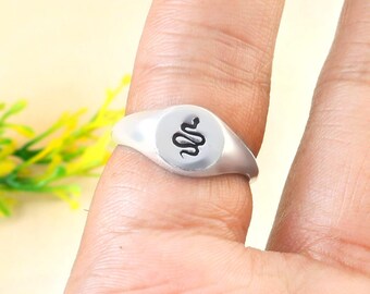 May include: A silver ring with a circular face featuring a black snake design. The ring is worn on a finger, showcasing its polished metal finish and the detailed snake illustration. The ring is a piece of jewelry.