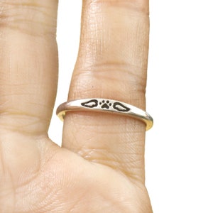May include: A silver ring with a paw print and two wings on it. The ring is worn on a person's finger.