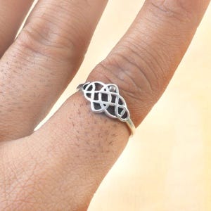 May include: A silver ring with a Celtic knot design. The ring features an openwork pattern of interwoven lines, creating a symmetrical, decorative shape. The ring is worn on a finger.