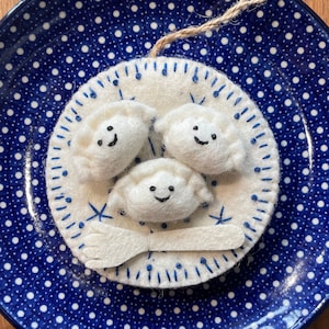 May include: Three white felt dumplings with smiling faces, stitched together with a white felt chopstick, on a blue and white polka dot plate.