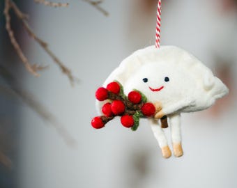 May include: A white felt dumpling ornament with a smiling face, black eyes, and a red stitched mouth. The figure holds a cluster of bright red berries on green stems and hangs from a red and white striped string against a blurred background.
