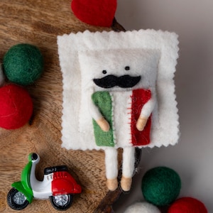 May include: A felt doll with a black mustache and an Italian flag design. The doll is surrounded by red, green, and white felt balls, a red heart, and a miniature scooter with an Italian flag design. The items are arranged on a wooden surface.