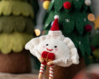 May include: A felted white dumpling ornament with a smiling face, wearing a small red Santa hat. The figure holds a tiny red gift box with gold ribbon and has striped red and white legs. The ornament is crafted from soft felt material.