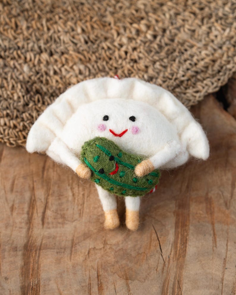 以下が含まれることがあります： 笑顔の顔、ピンクの頬、そして刺繍された顔を持つ緑色のフェルト製ピクルスを抱えた2本の小さな腕を持つ、白いフェルト製の餃子型オーナメント。餃子の上部はひだ状になっており、本体の下部からは2本の小さな足が伸びています。