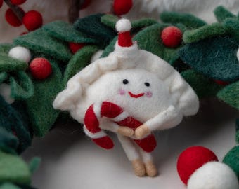 May include: A white felt dumpling ornament with a smiling face, pink cheeks, and a small red and white hat. The figure holds a red and white striped candy cane. It is positioned against a green felt wreath with red and white felt berries.