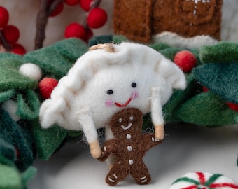 May include: A felt ornament featuring a white dumpling character with a smiling face holding a small brown gingerbread man. The ornament is surrounded by green felt leaves, red berries, a felt gingerbread house, and a round white peppermint candy.