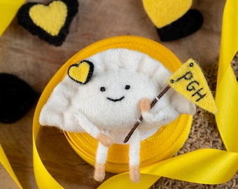 May include: A whimsical felt dumpling character with a smiling face and holding a small flag with "PGH" on it. The dumpling is surrounded by yellow ribbon and heart-shaped felt decorations in yellow and black.