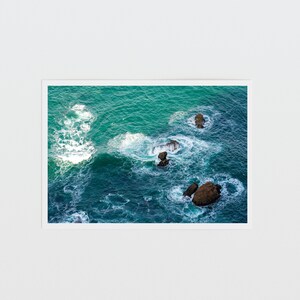 May include: An aerial view of a turquoise ocean with white foamy waves crashing against dark brown rocks.