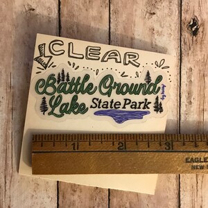 May include: A green and white sticker with the text "Clear Battle Ground Lake State Park" with a blue lake graphic. The sticker is on a wooden surface.