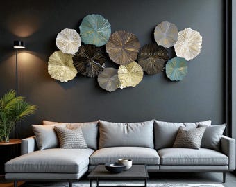 Op de afbeelding: Een moderne woonkamer met een grijze hoekbank, een zwarte salontafel en een patroonkleed. De muur is versierd met metalen, cirkelvormige sculpturen in goud, zilver en blauw. Een staande lamp en een plant in pot zijn ook aanwezig.