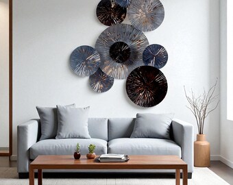 Op de afbeelding: Een moderne woonkamer met een grijze bank, een houten salontafel en een decoratieve wandkunst. De wandkunst bestaat uit meerdere metalen, cirkelvormige elementen in blauw, zilver en brons, die een opvallend middelpunt vormen.