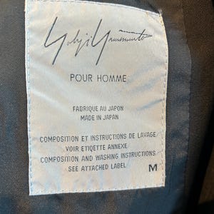 May include: A close-up of a label with the text "Yohji Yamamoto POUR HOMME" in black. The label is white with black text and the size is M. The label also says "Made in Japan".