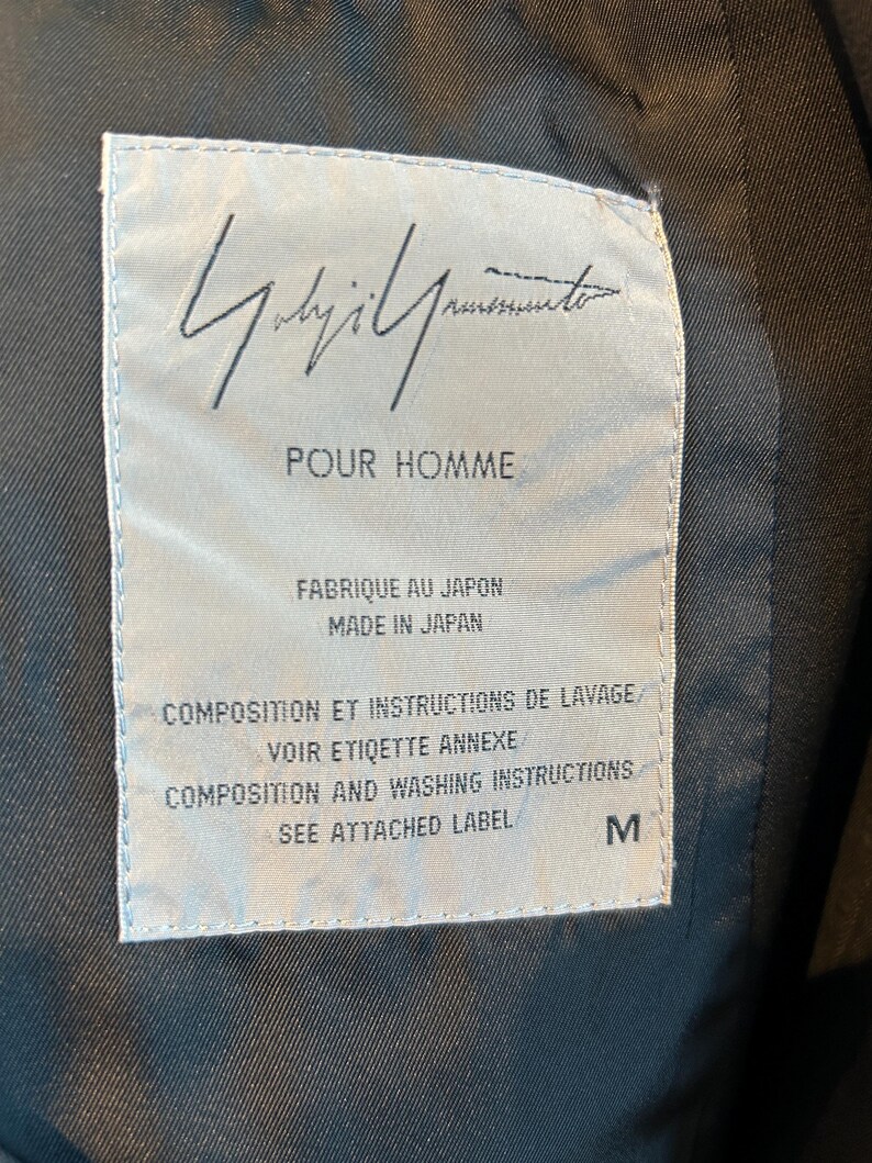 May include: A close-up of a label with the text "Yohji Yamamoto POUR HOMME" in black. The label is white with black text and the size is M. The label also says "Made in Japan".