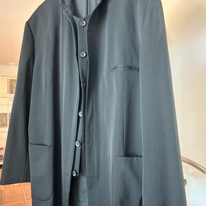 May include: A black suit jacket and matching vest hang on a wooden hanger. The jacket has two front pockets and a notched collar. The vest has three buttons. The suit is made of a smooth, dark fabric.