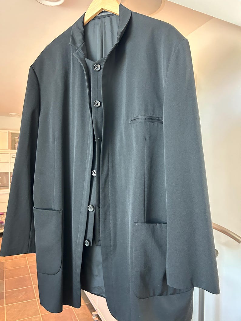 May include: A black suit jacket and matching vest hang on a wooden hanger. The jacket has two front pockets and a notched collar. The vest has three buttons. The suit is made of a smooth, dark fabric.