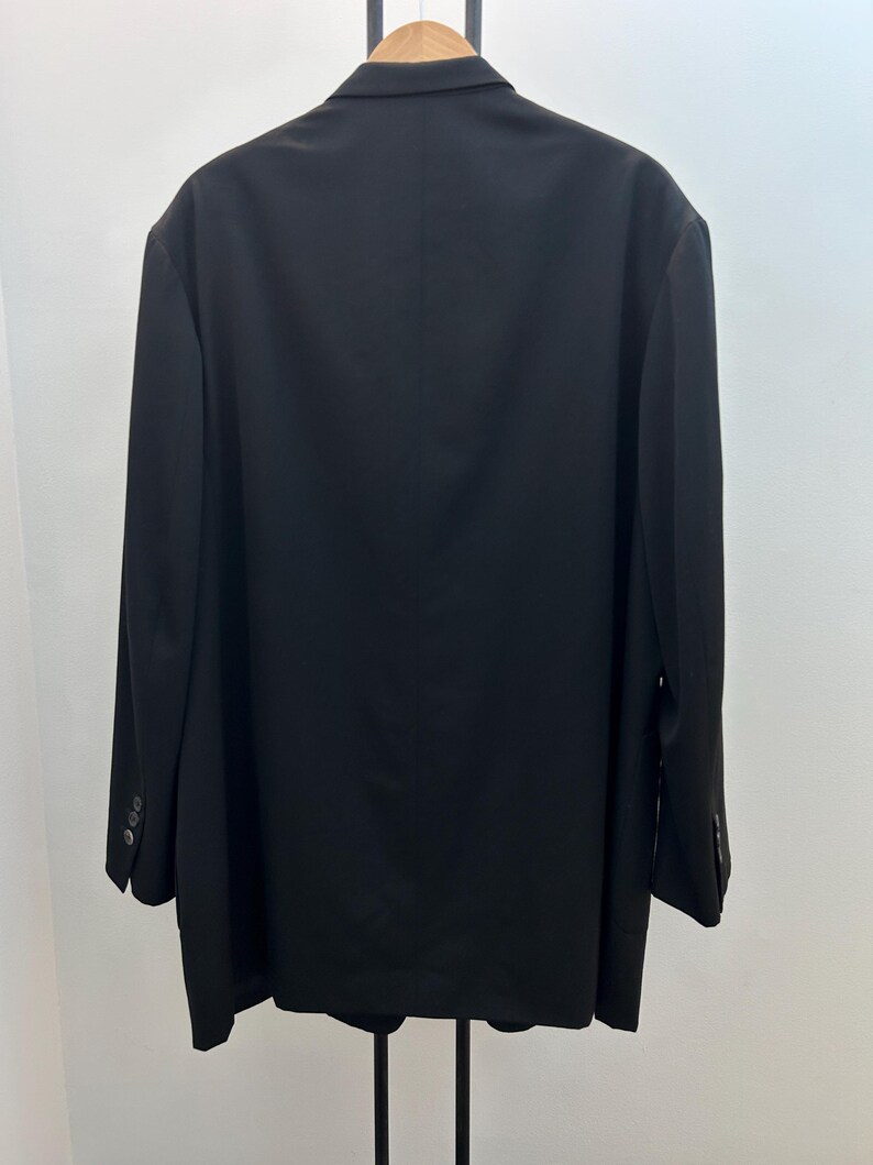 May include: A black blazer hanging on a wooden hanger. The blazer has a classic design with a notched lapel and three buttons on each cuff. The back of the blazer is smooth and tailored, with a single vent.
