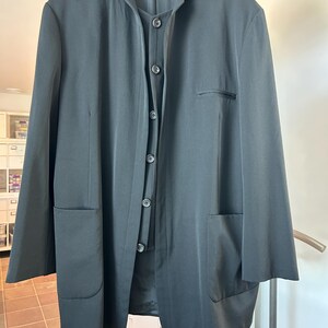 May include: Black long-sleeve jacket with a mandarin collar, button-up front, and two large front pockets. The jacket is hanging on a wooden hanger. A black button-up vest is visible underneath the jacket.