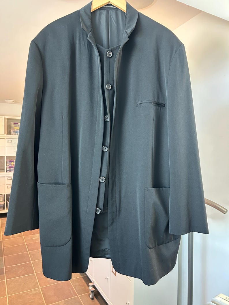 May include: Black long-sleeve jacket with a mandarin collar, button-up front, and two large front pockets. The jacket is hanging on a wooden hanger. A black button-up vest is visible underneath the jacket.