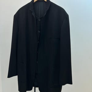 May include: A black blazer and vest set hanging on a wooden hanger. The blazer has a mandarin collar, long sleeves, and two front pockets. The vest is buttoned up. The set is displayed against a white background.