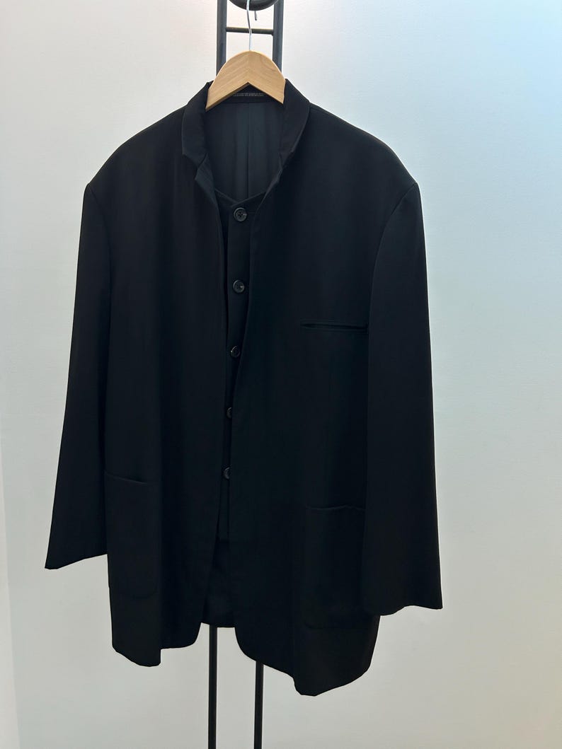 May include: A black blazer and vest set hanging on a wooden hanger. The blazer has a mandarin collar, long sleeves, and two front pockets. The vest is buttoned up. The set is displayed against a white background.