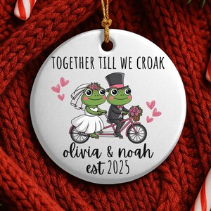 Peut inclure: Ornement de Noël rond en céramique blanche avec une ficelle dorée. Il représente un couple de grenouilles de dessin animé à vélo, avec le texte "TOGETHER TILL WE CROAK", les noms "olivia & noah" et l'année "est 2025". Des cœurs et des fleurs roses sont également présents.