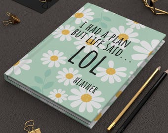 May include: A journal with a light green cover featuring white daisies with yellow centers. The cover reads "I HAD A PLAN BUT LIFE SAID... LOL HEATHER". Gold and black pencils and gold binder clips are also visible.