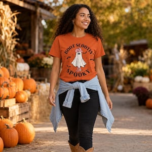 May include: An orange short-sleeve t-shirt featuring a graphic of a ghost wearing a pink cowboy hat and boots, surrounded by small bats and the text "BOOT SCOOTIN' SPOOKY". A light blue denim shirt is tied around the waist over black pants.