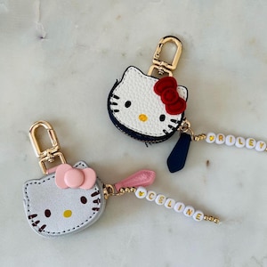 May include: Two Hello Kitty-shaped keychains. One is white with a red bow, the other is gray with a pink bow. Both have gold-colored hardware and beaded name tags that spell out "RILEY" and "CELINE".