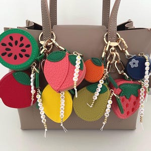 May include: A collection of colorful fruit-shaped keychains, including a watermelon, apple, peach, orange, lemon, avocado, strawberry, and blueberry. Each keychain has a gold-colored chain and a beaded name tag. The keychains are attached to a taupe handbag.