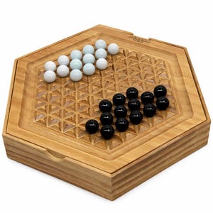 May include: Wooden board game with a hexagonal shape, featuring a carved geometric pattern. The game includes white and black marbles. The game board is made of light-colored wood, and the marbles are arranged on the playing surface.