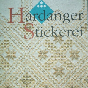 Op de afbeelding: Een boekomslag met de titel "Hardanger Stickerei" in rode en zwarte tekst. De omslag heeft een ruitvormig ontwerp met een blauwe achtergrond en een witte rand. De achtergrond van de omslag is een beige stof met een wit kantpatroon.