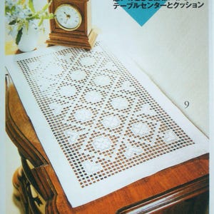 May include: A white rectangular crochet table runner with a repeating geometric grid pattern featuring floral-like motifs. The runner is displayed on a wooden surface next to a wooden clock and a vase of flowers, with Japanese text visible.