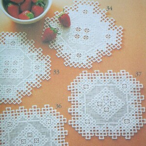 May include: Four white square doilies featuring intricate cutwork and openwork geometric patterns, labeled with the numbers 33, 34, 36, and 37. A white bowl filled with fresh red strawberries is positioned in the upper left corner of the frame.