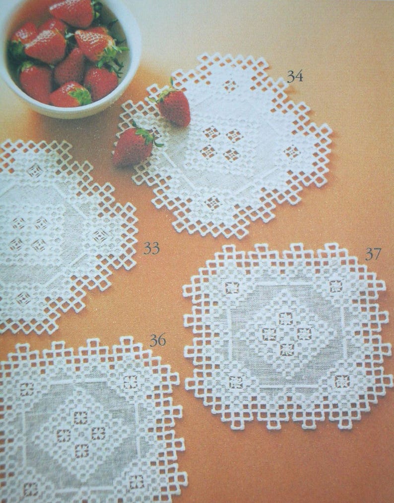 May include: Four white square doilies featuring intricate cutwork and openwork geometric patterns, labeled with the numbers 33, 34, 36, and 37. A white bowl filled with fresh red strawberries is positioned in the upper left corner of the frame.