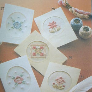 May include: Five cream-colored greeting cards featuring embroidered floral and geometric designs within circular or oval cutouts. The cards are numbered 27, 28, 29, and 31. Included are two small spools of thread and a length of twisted embroidery floss.