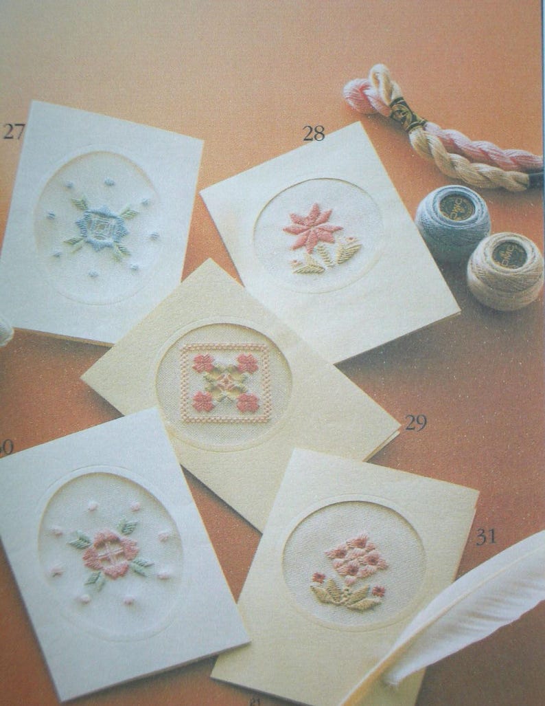 May include: Five cream-colored greeting cards featuring embroidered floral and geometric designs within circular or oval cutouts. The cards are numbered 27, 28, 29, and 31. Included are two small spools of thread and a length of twisted embroidery floss.
