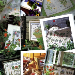 May include: A collection of cross-stitch embroidery designs featuring various floral and garden themes. The designs include a teacup with flowers, a basket of flowers, a garden scene with a house, and a variety of individual flowers. The designs are framed and displayed on a table and wall.