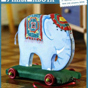 Op de afbeelding: Een blauw houten olifanten speelgoed met een kleurrijk, gepatroneerd zadel, op een groene stoffen basis met rode wielen. Het speelgoed staat op een houten vloer. De tekst op de tijdschriftcover luidt "РУЧНАЯ РАБОТА" en "авторская бумага и чехлы на стулья" met het nummer en de datum van de uitgave.