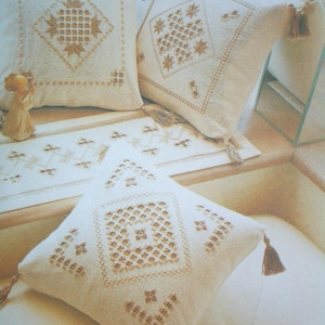 May include: Three square off-white throw pillows and a matching rectangular table runner, each featuring intricate tan embroidery in geometric diamond patterns. The pillows are finished with small corner tassels and labeled with the numbers 6, 7, 8, and 9.