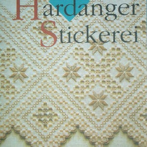 May include: A cream-colored fabric featuring intricate Hardanger embroidery with geometric diamond patterns, star-shaped motifs, and a scalloped edge. The text Hardanger Stickerei is printed in dark lettering over a blue diamond-shaped graphic.
