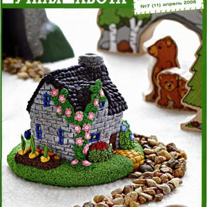 May include: A detailed miniature stone house with a black tiled roof, decorated with pink flowers and green foliage. It's set on a green base with a garden and a stone path. The image is from a magazine cover with Russian text.