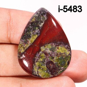 Puede incluir: Un cabochón de piedra preciosa roja, verde y morada con forma de lágrima. La piedra tiene un patrón único de colores en espiral y está pulida a un acabado liso.