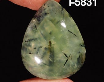 May include: A green and black gemstone cabochon with a teardrop shape. The stone has a translucent appearance with black inclusions throughout. The stone is labeled "i-5831".