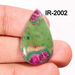Peut inclure: Un cabochon de pierre précieuse vert et rose en forme de larme. La pierre a une base verte avec des inclusions roses et rouges. La pierre est étiquetée "IR-2002".